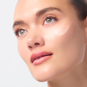 Close-up of a woman's face with a glossy, dewy complexion showing a liquid beauty product applied to her cheek against a light gray background.